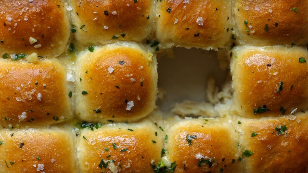 Golden-brown garlic vegan dinner rolls arranged in a grid with one missing piece, topped with herbs and sea salt, showcasing a soft, fluffy interior from a birds-eye view.